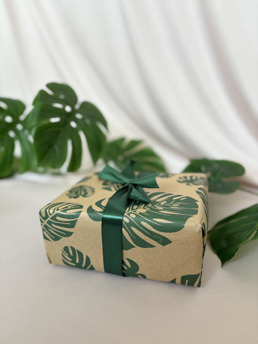 Gift box wrapped in paper with leaf pattern and green ribbon on a white surface with green leaves in the background