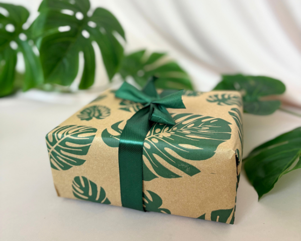 Gift box wrapped in brown paper with green leaf pattern and green ribbon, placed on a surface with green leaves in the background.