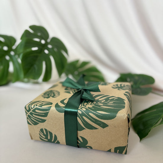 Gift box wrapped in paper with leaf pattern and green ribbon on a white surface with green leaves in the background