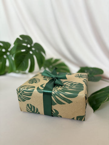 Gift box wrapped in paper with leaf pattern and green ribbon on a white surface with green leaves in the background