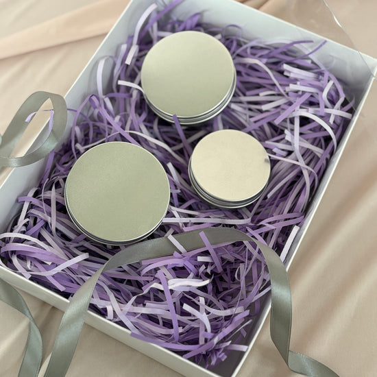 Three small round containers with lids in a box filled with purple shredded paper on a beige surface.
