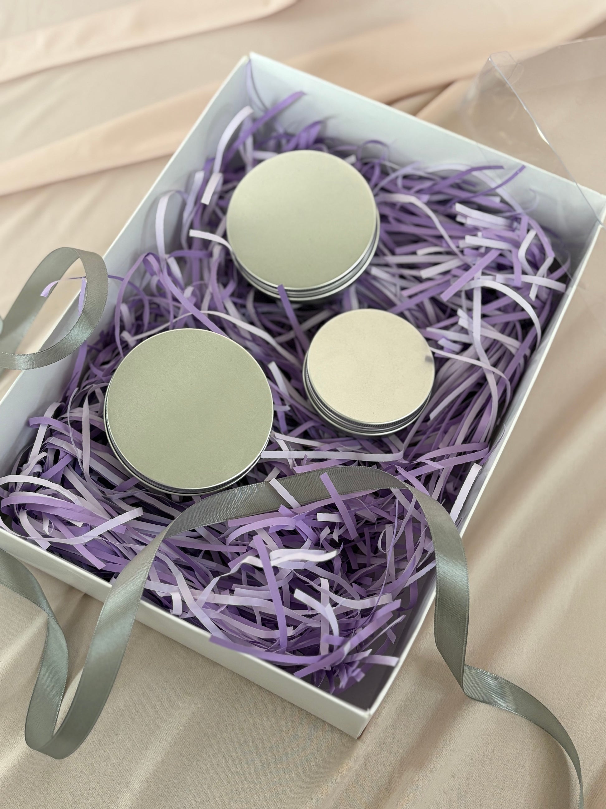 Three small round containers with lids in a box filled with purple shredded paper on a beige surface.