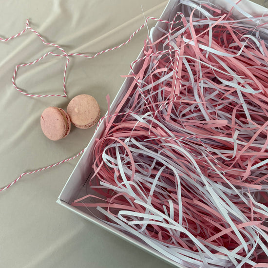 Box of pink and white shredded paper with two macarons on a light background